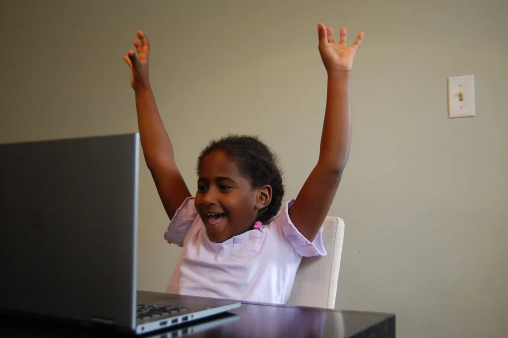 student holding her hand up, smiling, and looking at a computer, where she's in the midst of a powered by reading partners tutoring session