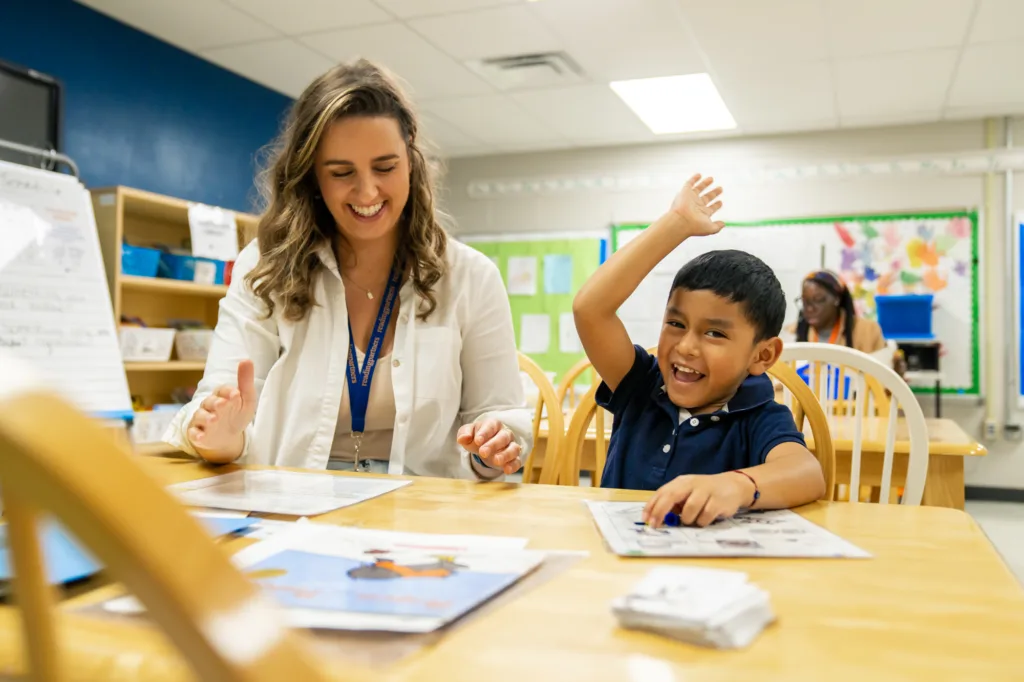 student with his hand in the air, smiling widely while sitting next to his tutor