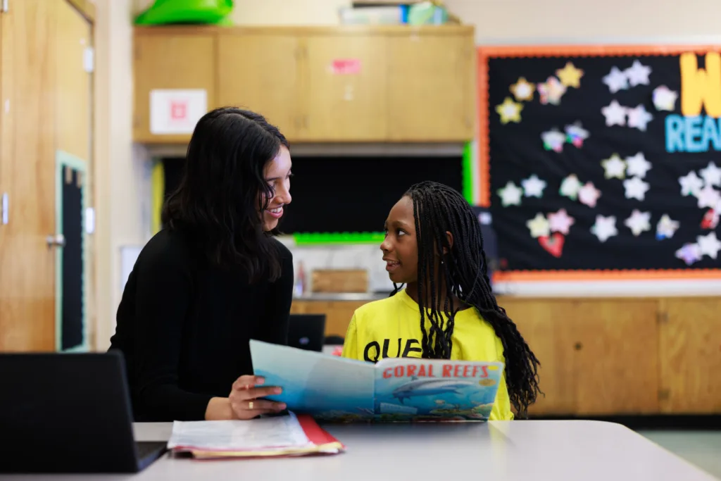 a student and tutor sit with a book in hand, smiling at each other
